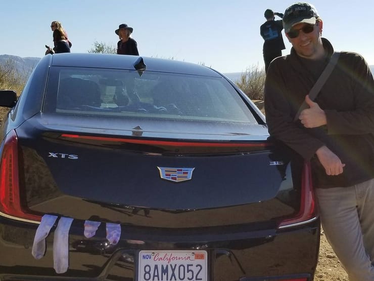 Dan giving a thumbs-up beside a black Cadillac on a dusty hillside pull-off, socks hung over the boot lid to dry