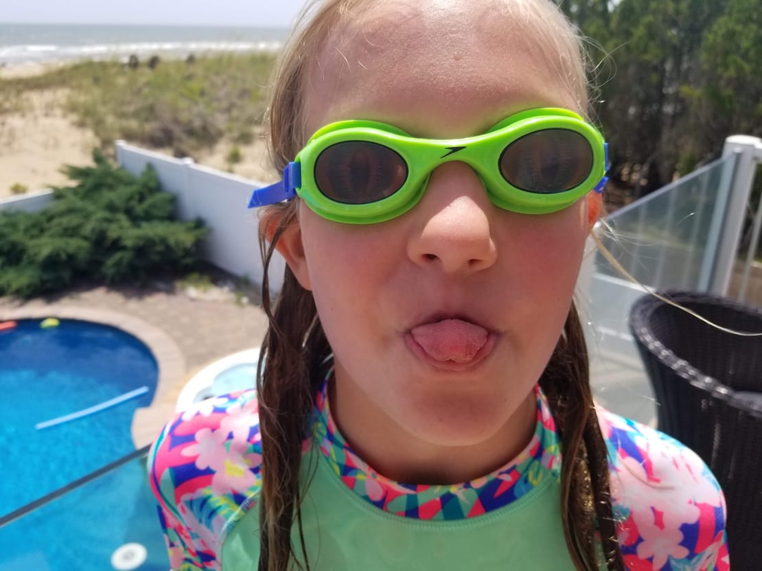 Evelyn in bright green swim goggles, poking her tongue out at the camera, with a pool and the ocean behind her